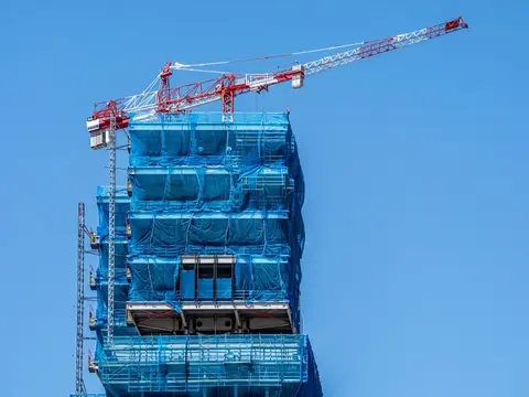 A red and white tower crane over a tall building wrapped in blue netting