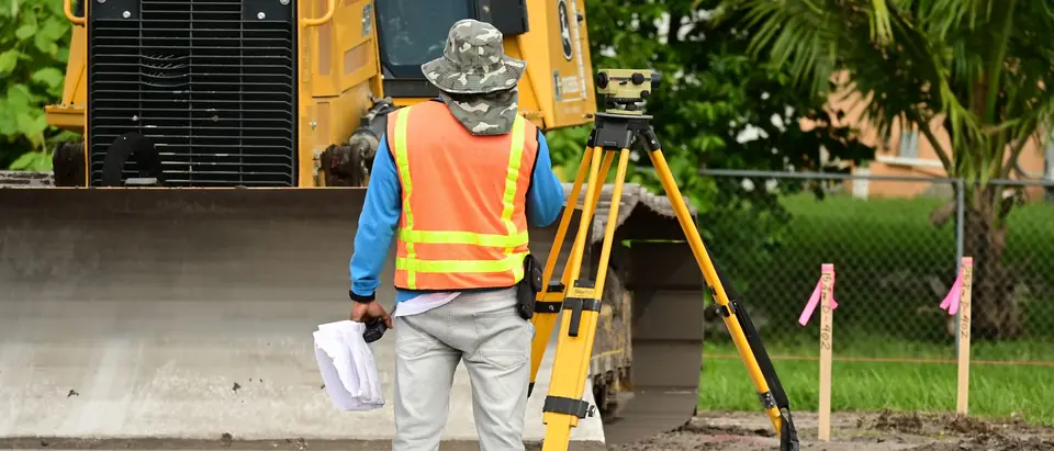 A surveyor setting up a tripod beside an excavator on a sunny site
