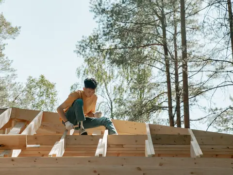 A builder working on the wood frame of a new building among tall trees