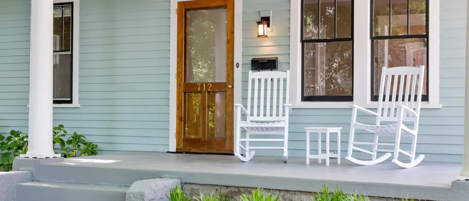 A pale green front porch with a wood door and white rocking chairs