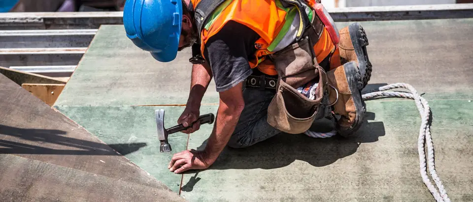A roofer in a blue hard hat and hi-vis vest nailing down roof decking
