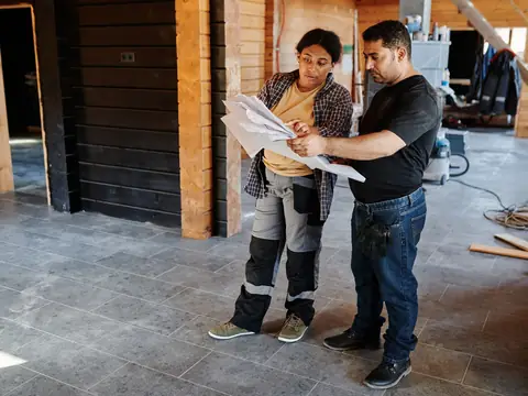 Two builders reading a printed plan inside a house being renovated