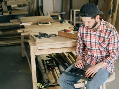 A woodworker using a laptop at his bench among stacks of lumber